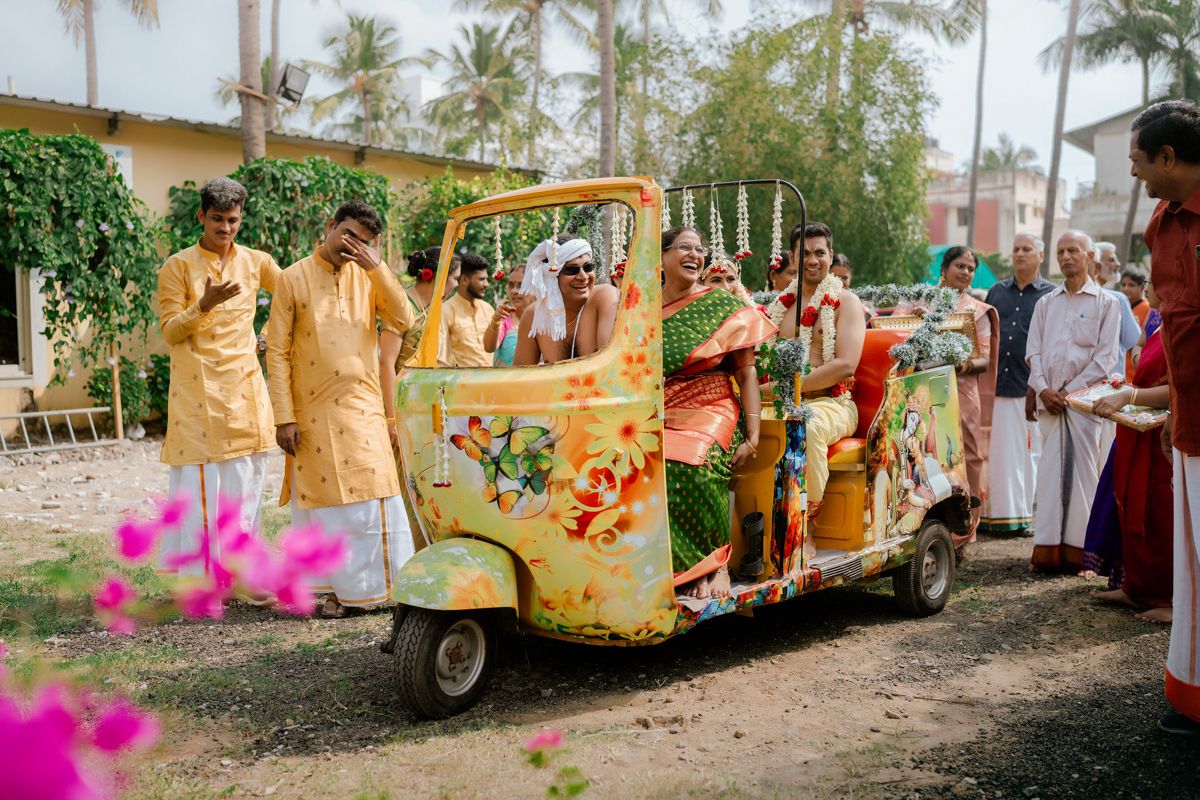 Decorated golf cart with cheerful wedding party in traditional Indian attire driving through palm-lined path.