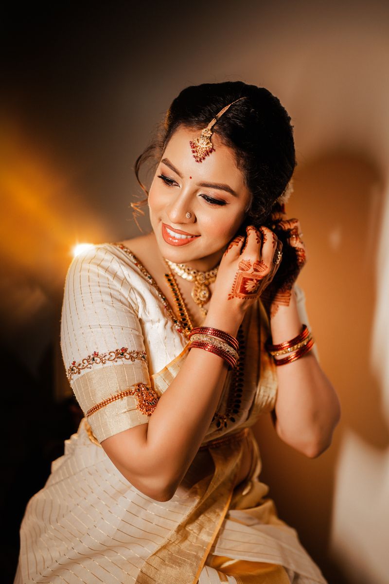 Traditional South Asian bride in white and gold saree with henna designs and jewelry poses elegantly in warm lighting.