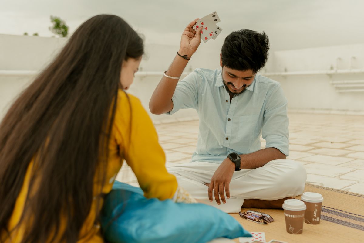 A couple playing cards while sitting on a rooftop, with the woman in yellow and man in light blue attire.