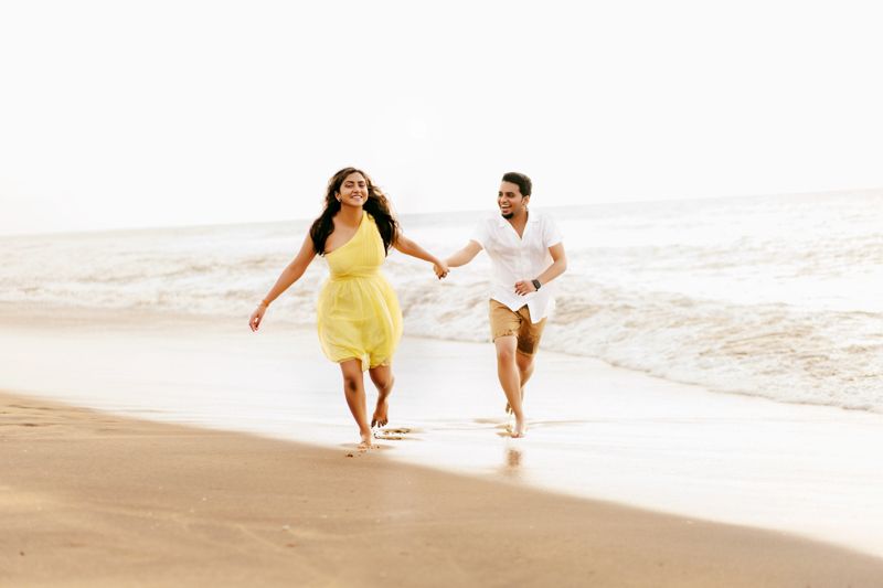 Couple running and holding hands along the beach shoreline, with woman in yellow dress and man in white.