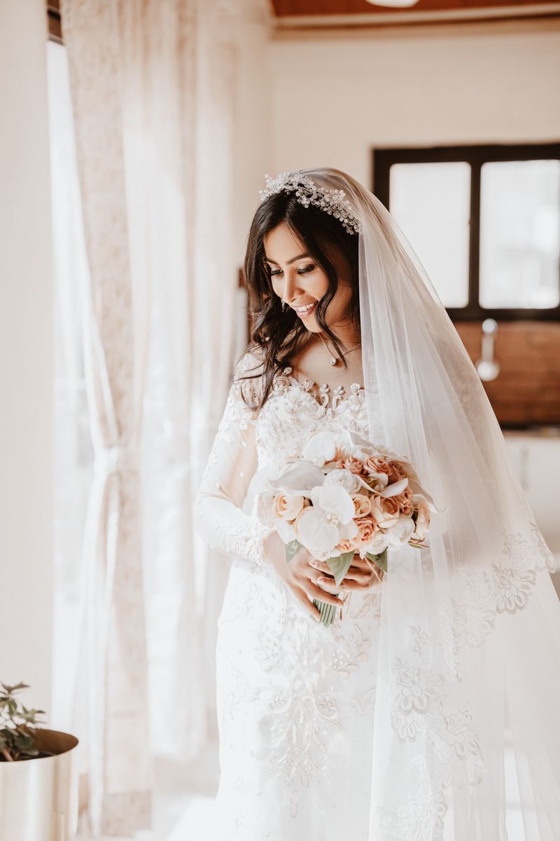 A bride in an elegant lace wedding gown with veil holds a delicate pink and white bouquet near a sunlit window.