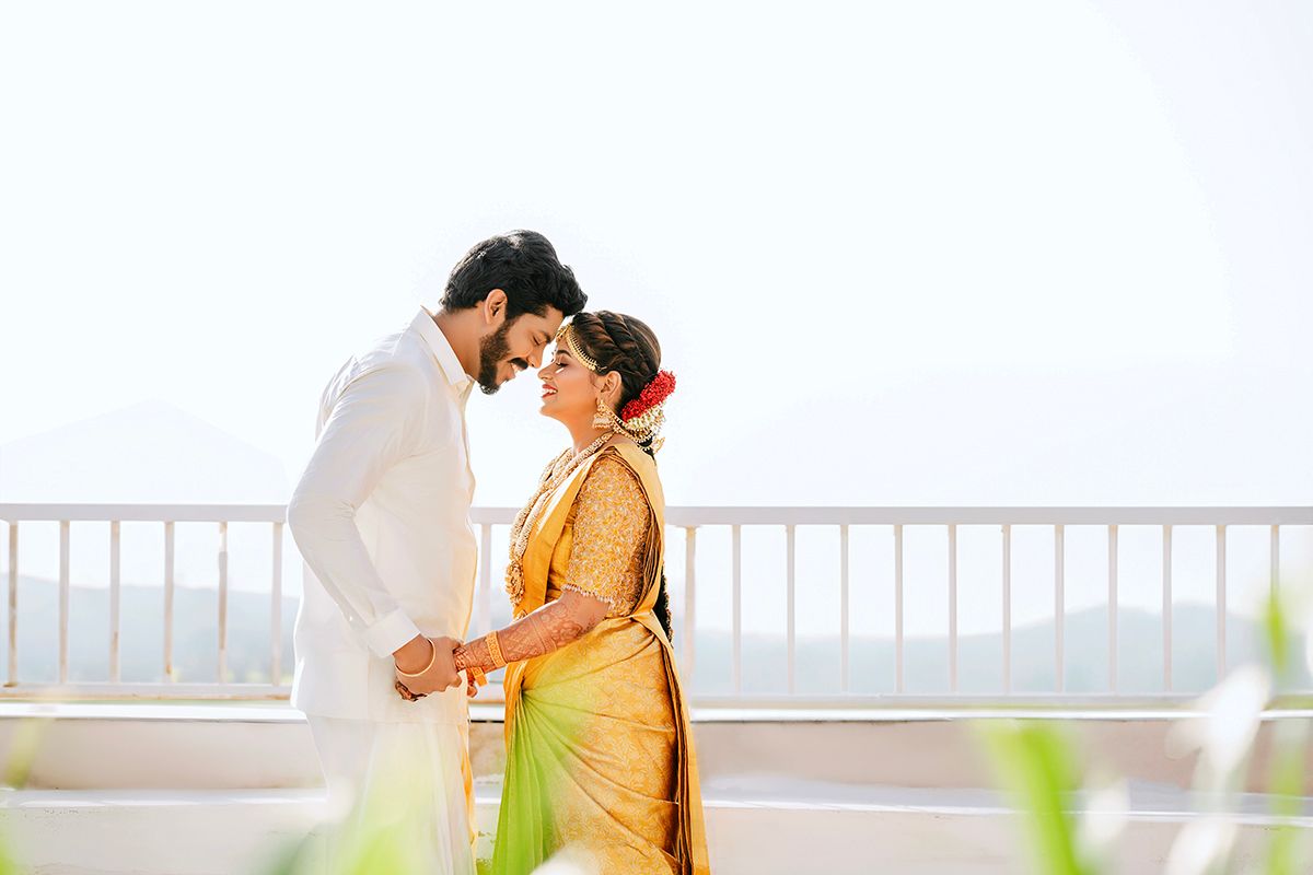 A romantic couple in traditional Indian attire share a tender moment on a balcony overlooking a misty landscape.