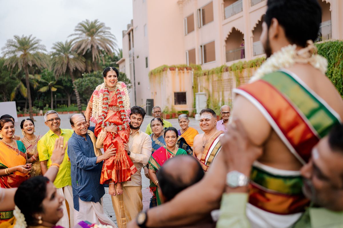 A festive group in traditional Indian wedding attire dances and celebrates during an outdoor ceremony at a venue with palm trees.