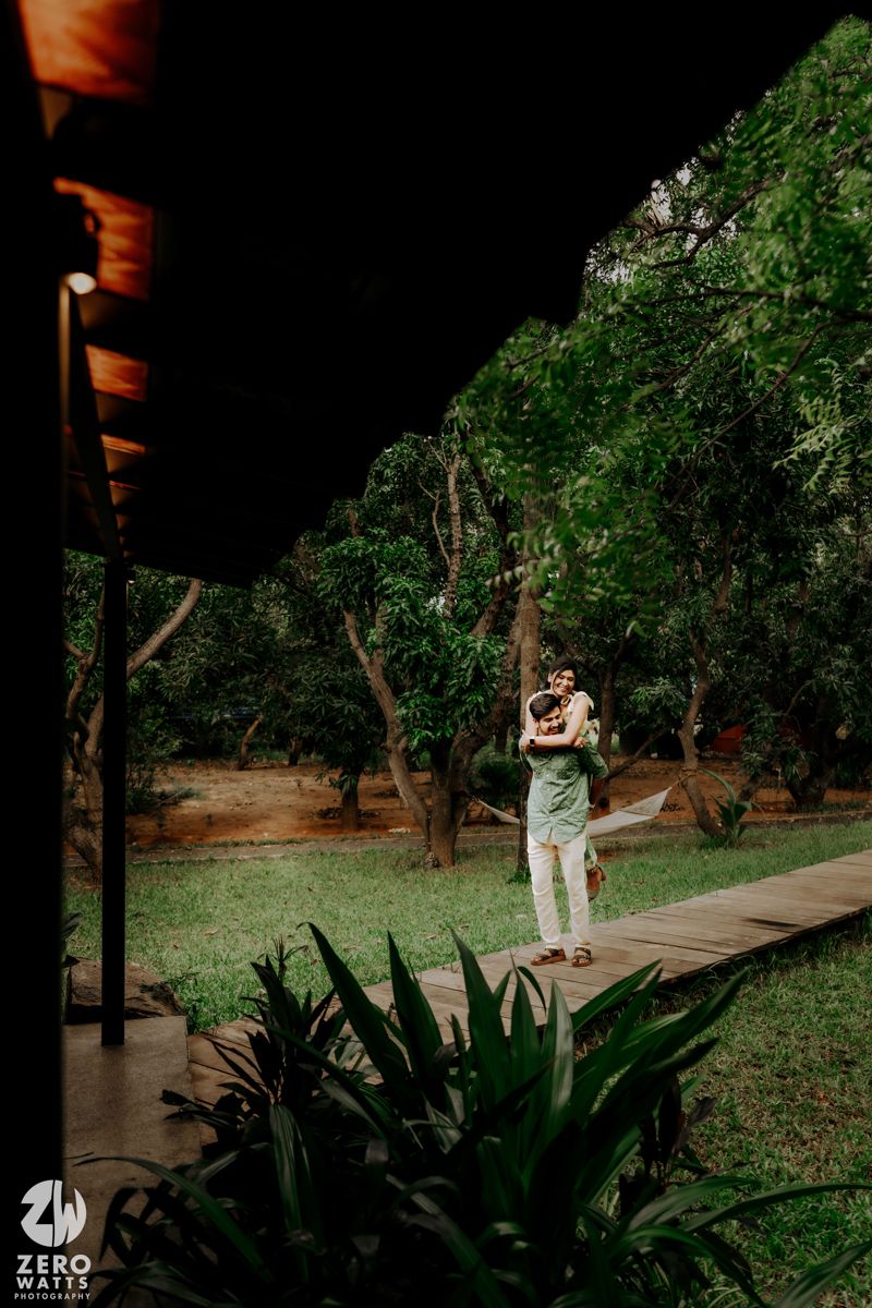 Figure in a light outfit stands on a wooden deck surrounded by lush greenery and tropical plants in a garden setting.
