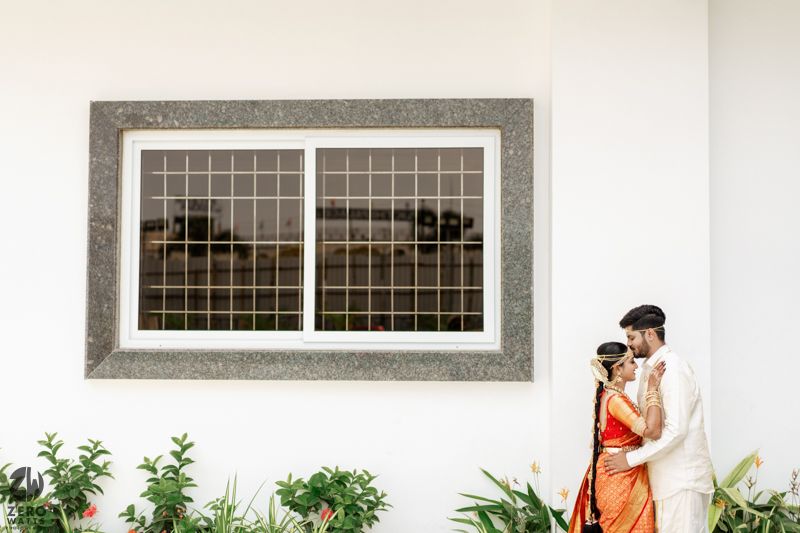 Couple in traditional Indian attire embrace near a white wall with a window and tropical plants.
