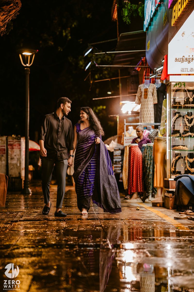 A couple walking on wet streets at night with neon lights and shop reflections in puddles.