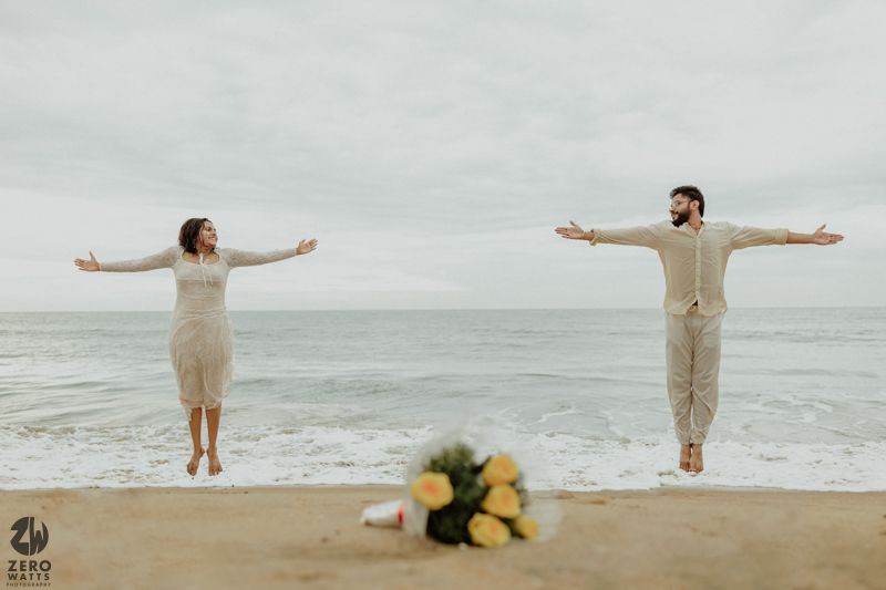 Two people in white outfits spread their arms joyfully on a serene beach.