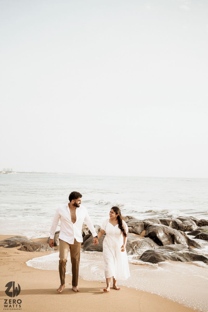Couple walking hand in hand along a rocky beach shoreline wearing white outfits.