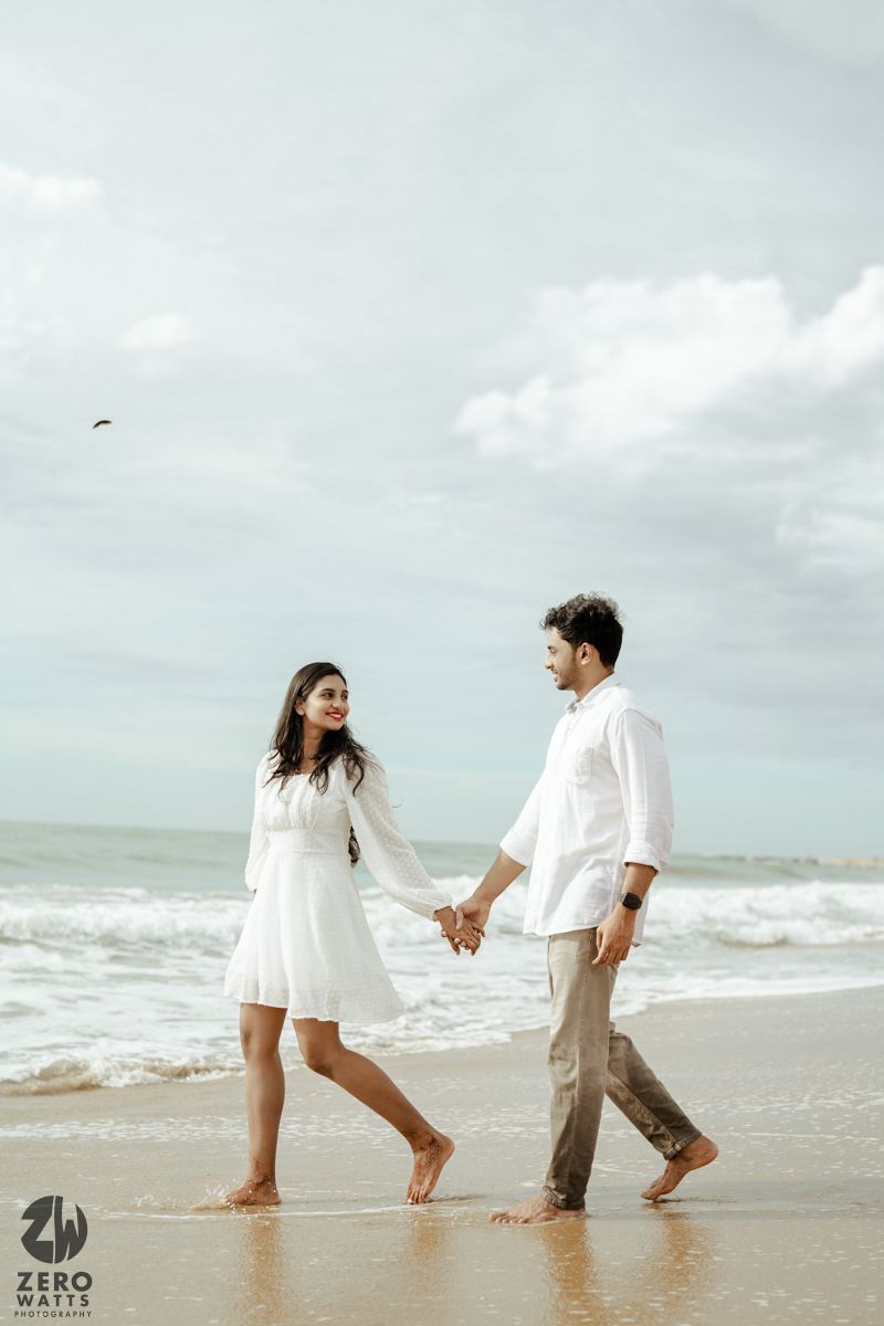 Couple in white clothing walks hand in hand along a beach shoreline on a cloudy day.