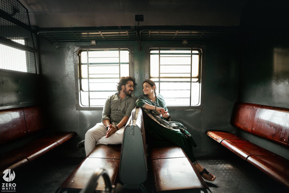 A couple sits facing each other on red benches inside a rustic train compartment with natural lighting.