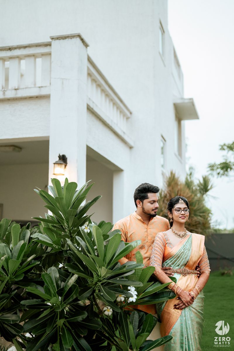 A couple in traditional South Asian attire pose together by lush green plants near a white building exterior.
