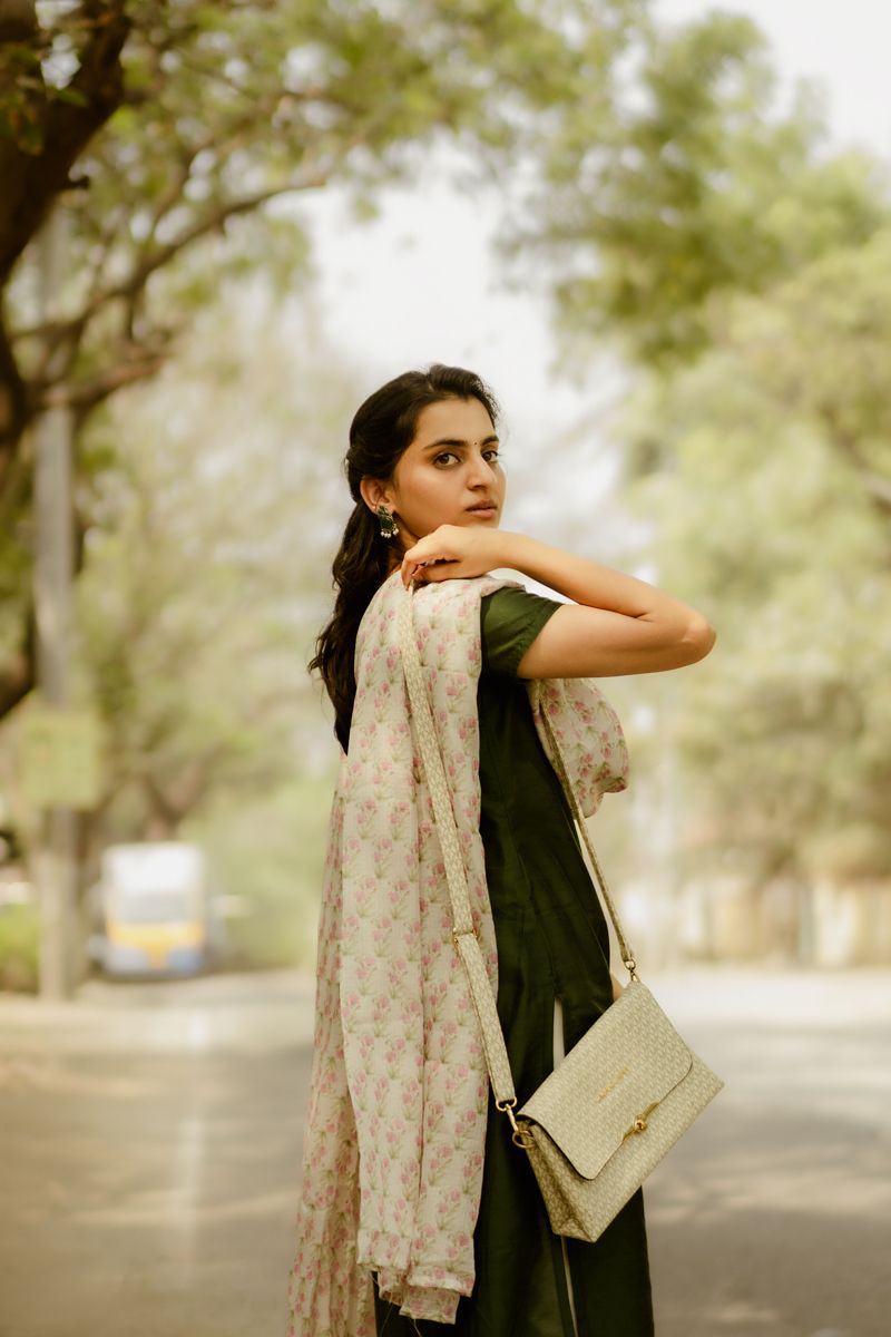 A woman in traditional Indian attire poses with a beige handbag against a blurred outdoor background.