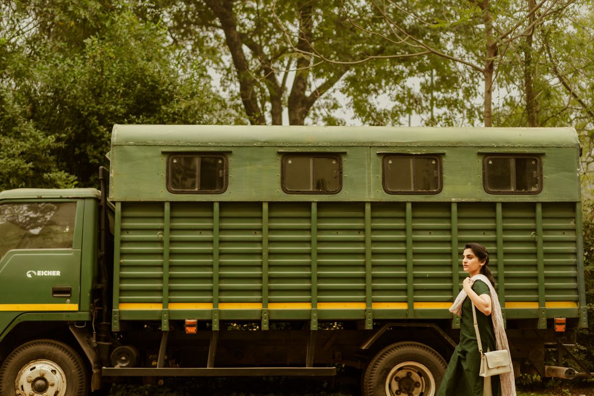 A large green transport truck parked under trees with someone standing beside it.