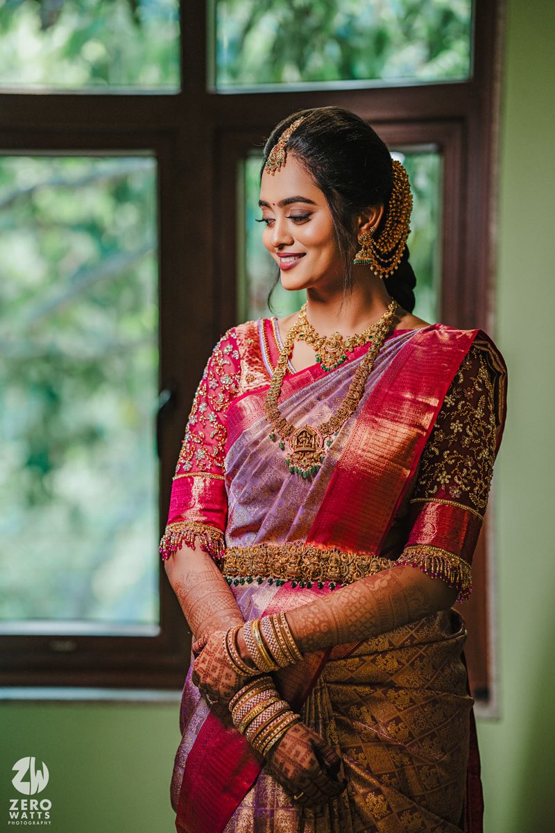 Traditional South Indian bridal portrait showing intricate jewelry and embroidered silk saree by a window.