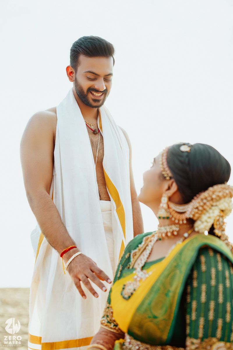 Wedding couple in traditional South Indian attire share a candid joyful moment against bright sunlit backdrop.