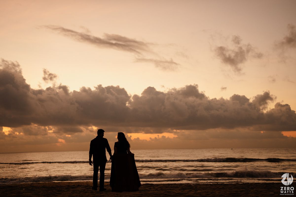 A silhouetted couple stands on the beach at sunset with dramatic orange clouds overhead.