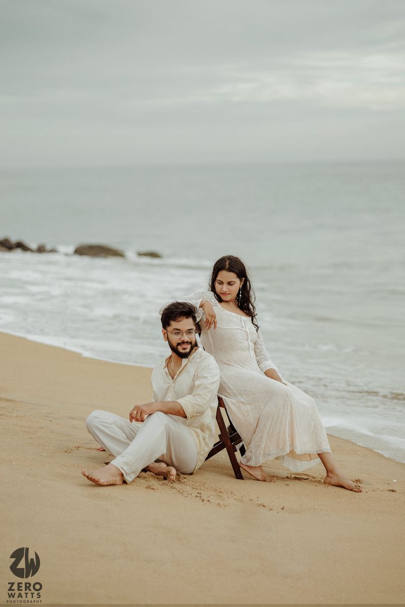 A couple in white outfits posing on a cloudy beach.