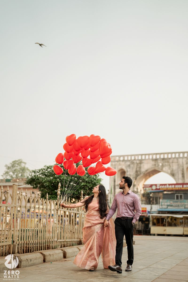 A couple holding red balloons walks along a promenade with historic architecture in the background.