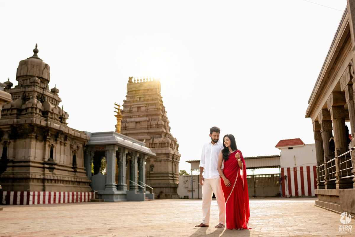 Couple in white and red attire poses at a traditional South Indian temple during golden hour sunset.