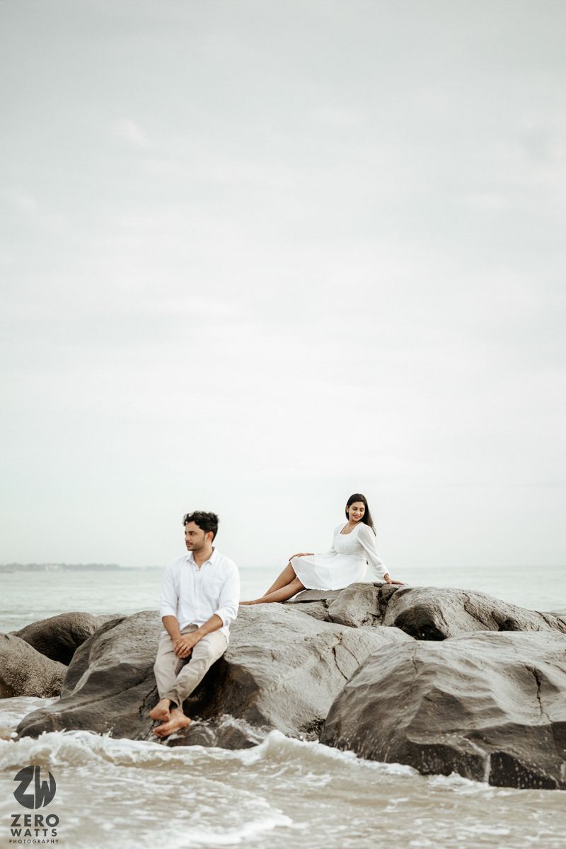 A couple in white sits separately on large coastal rocks with ocean waves below.