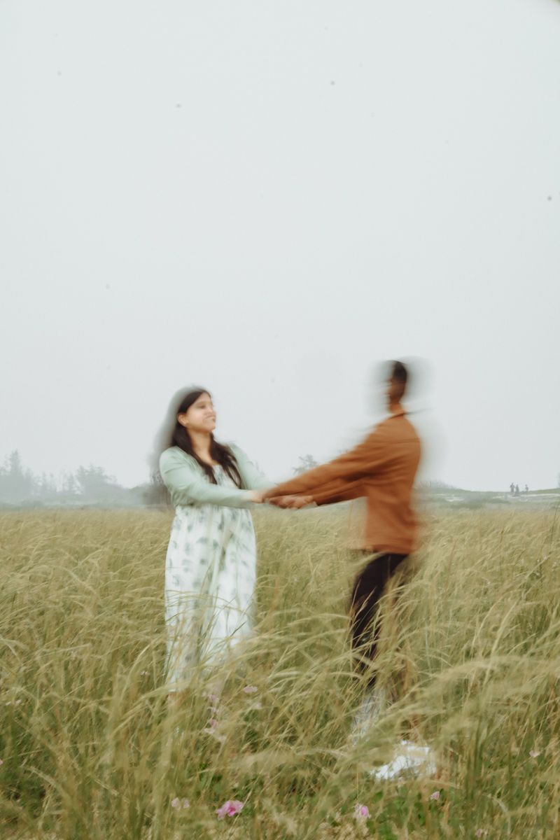 A couple dancing and spinning playfully together in a grassy meadow on an overcast day.