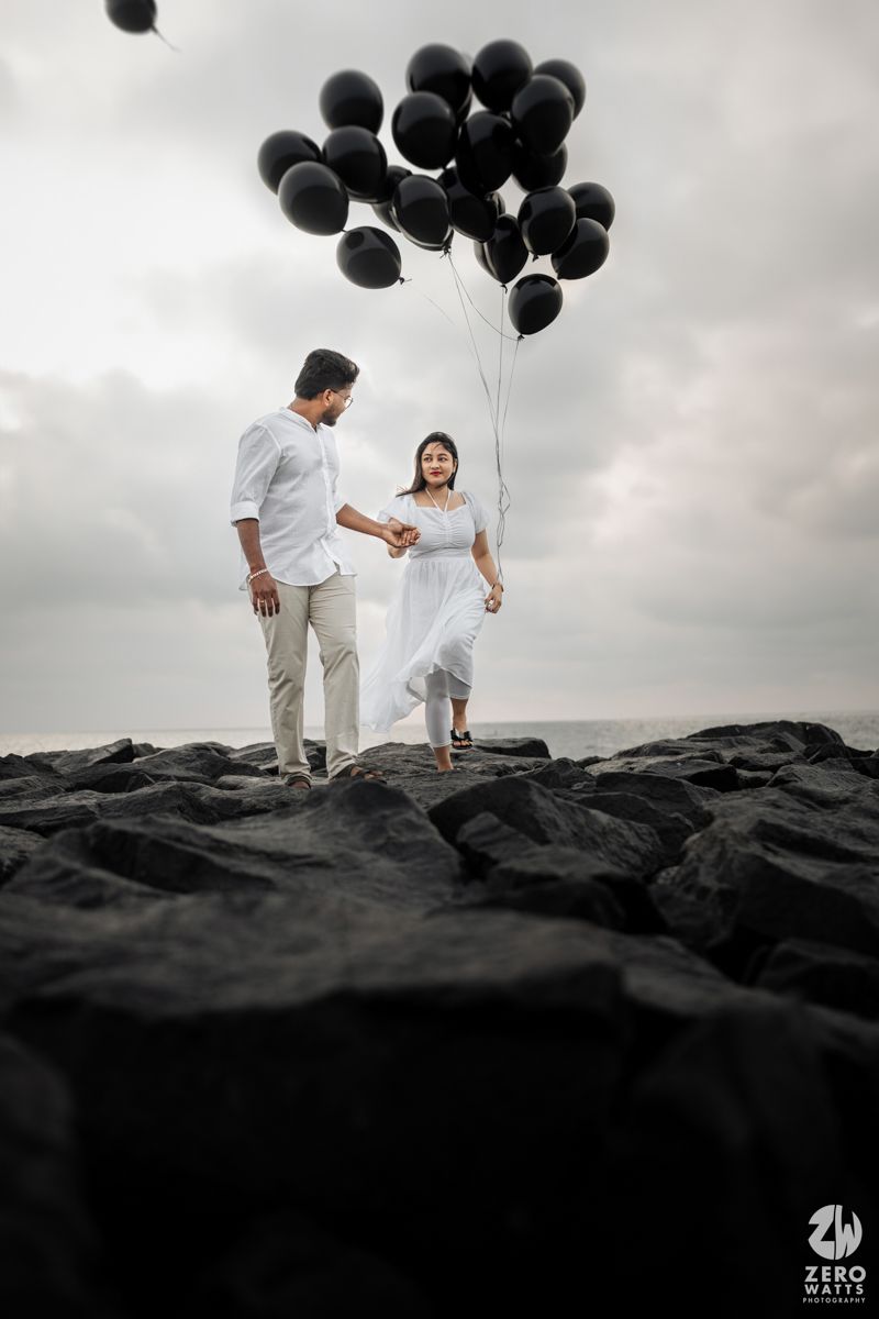 Couple with black balloons pose on rocky beach under cloudy sky.