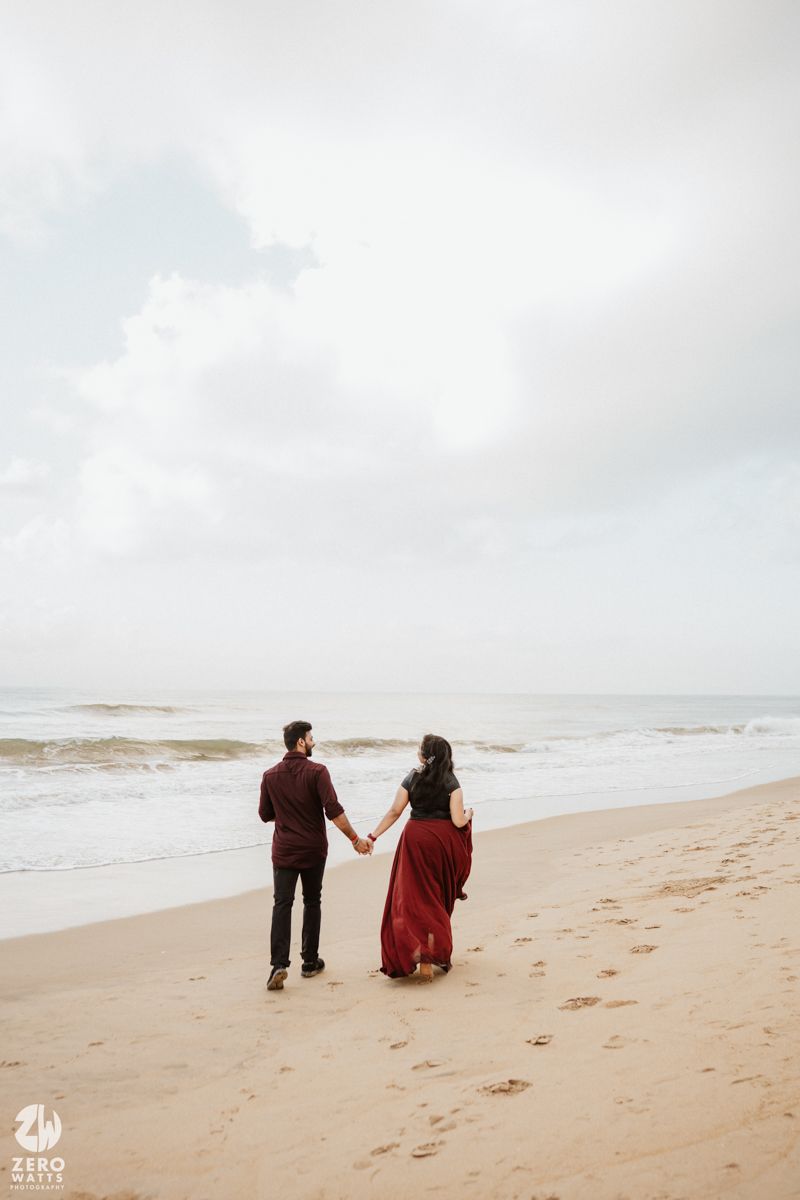 Couple walking hand in hand on a cloudy beach with waves crashing in the background.