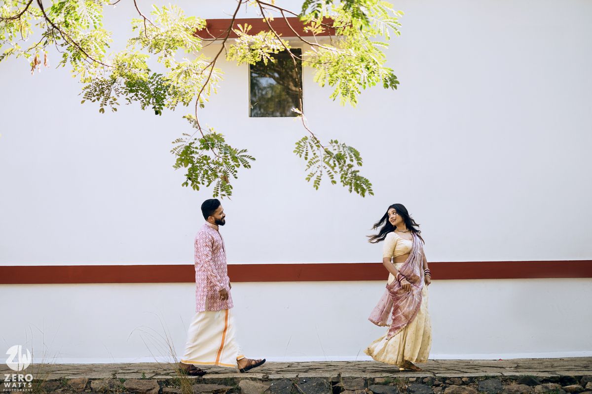 Two people in traditional attire walking by a white wall under green foliage.