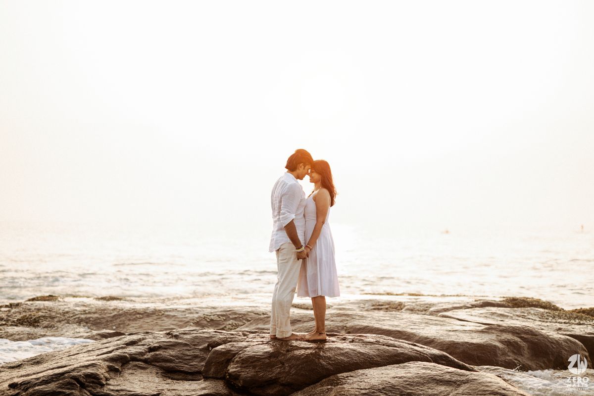 A romantic couple embraces while standing on coastal rocks during sunset at the beach.