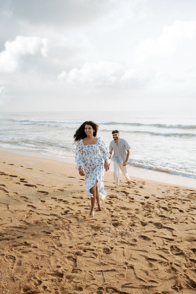 A couple runs playfully along a sandy beach with waves and cloudy skies in the background.