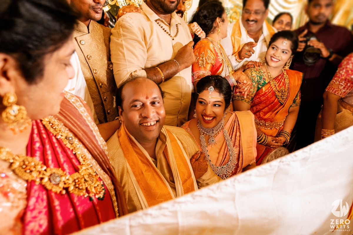 South Indian wedding ceremony with guests in traditional orange and red attire smiling during a ritual.