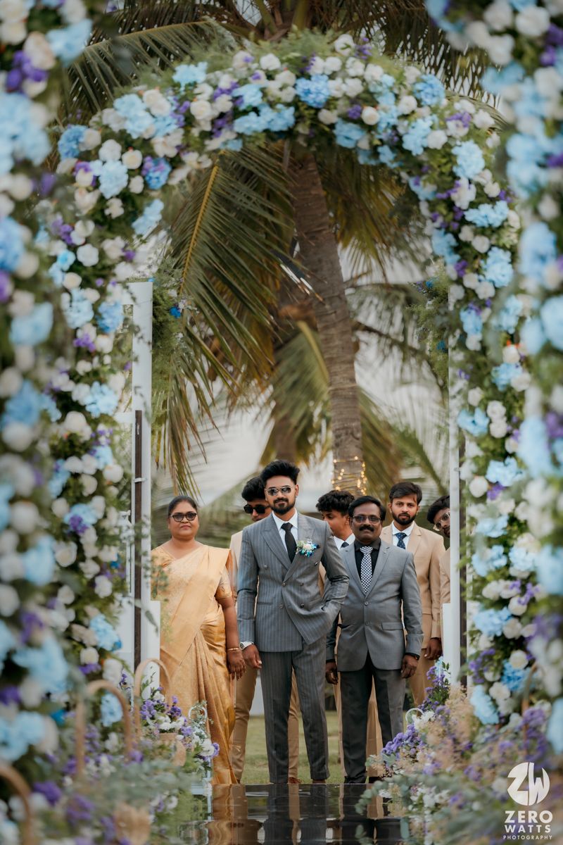 Wedding ceremony under a floral arch with blue and white blossoms against a tropical palm tree backdrop.