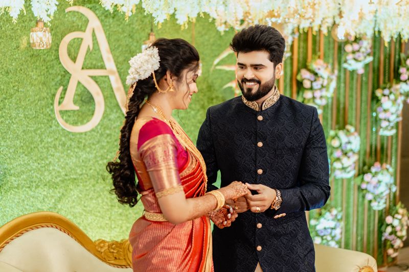 A couple at an engagement ceremony stands in front of a green backdrop with floral decorations.