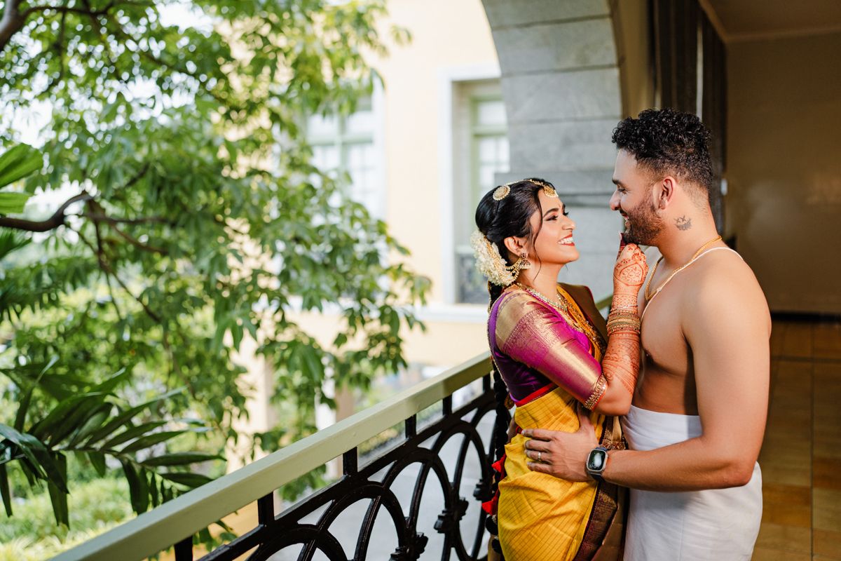 A romantic couple embraces on a balcony, wearing traditional South Asian attire with lush greenery in background.