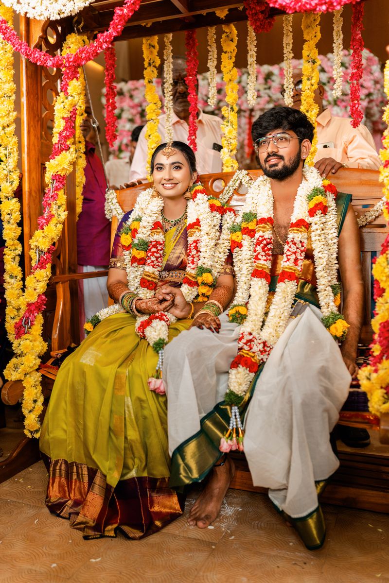 Newlyweds wearing traditional South Indian wedding attire and garlands seated together under decorated mandap.