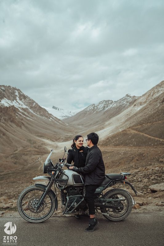 Two riders share an intimate moment on their motorcycle against a backdrop of snow-capped mountains on a cloudy day.