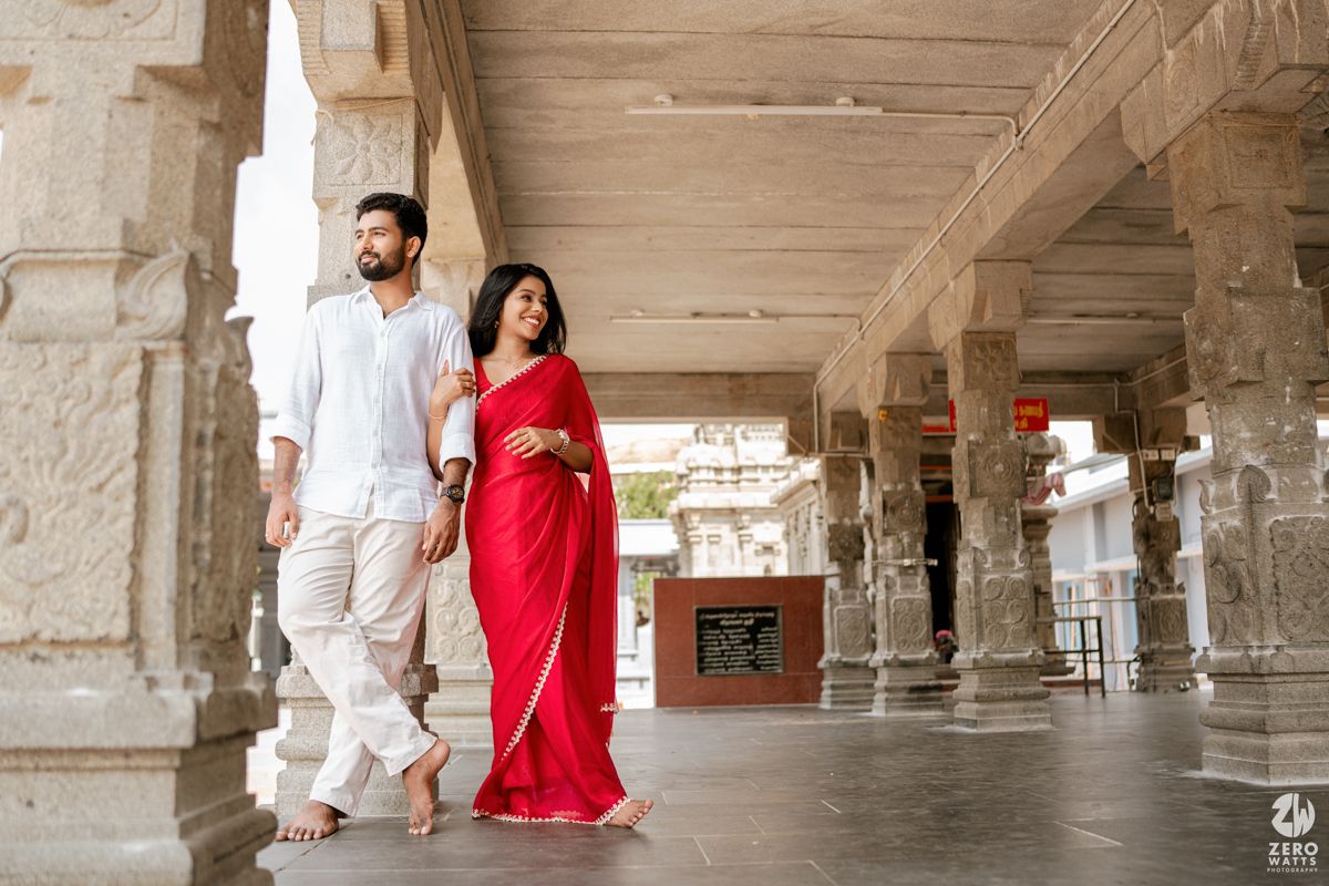 A couple walks hand in hand through a stone pillared corridor, with the woman wearing a vibrant red saree.