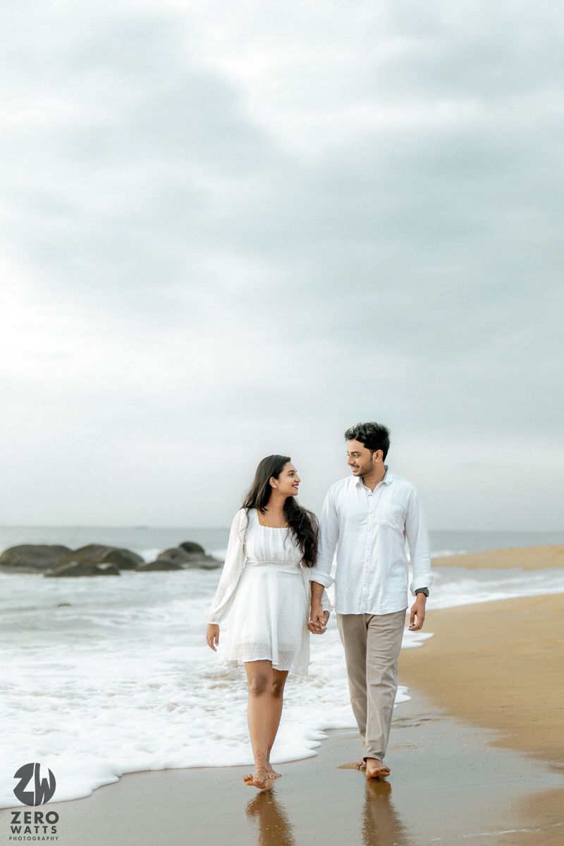 A couple in white outfits walks hand in hand along a scenic beach shore with waves crashing in the background.