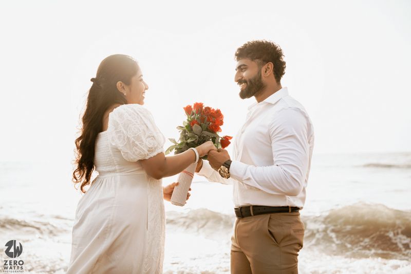 A couple shares a joyful moment with red flowers during their beach photoshoot.