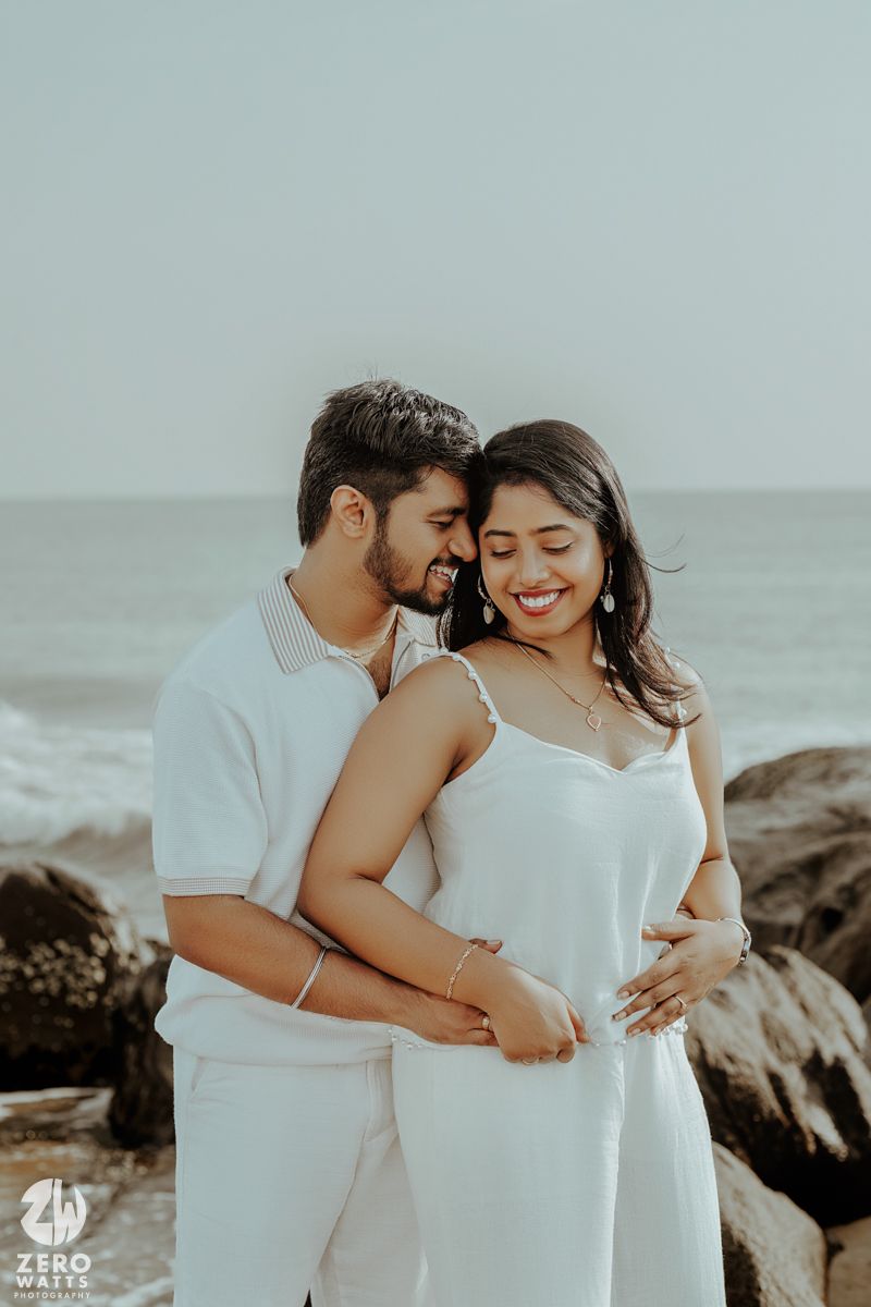 A romantic moment shared between a couple in white outfits on a rocky beach.
