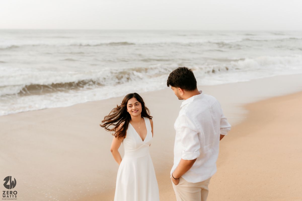 A couple in white clothing share a candid moment on a windswept beach.