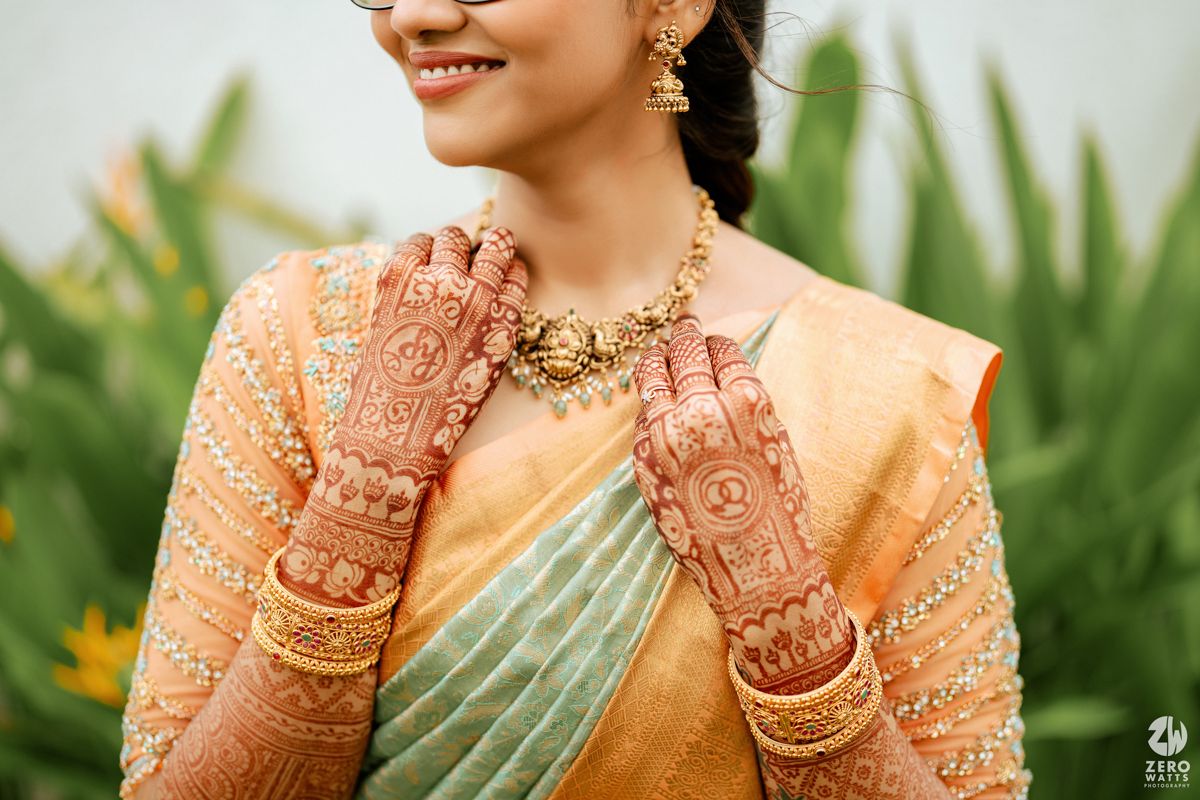 Traditional mehndi-adorned hands with intricate henna designs shown with gold jewelry and peach-colored saree.