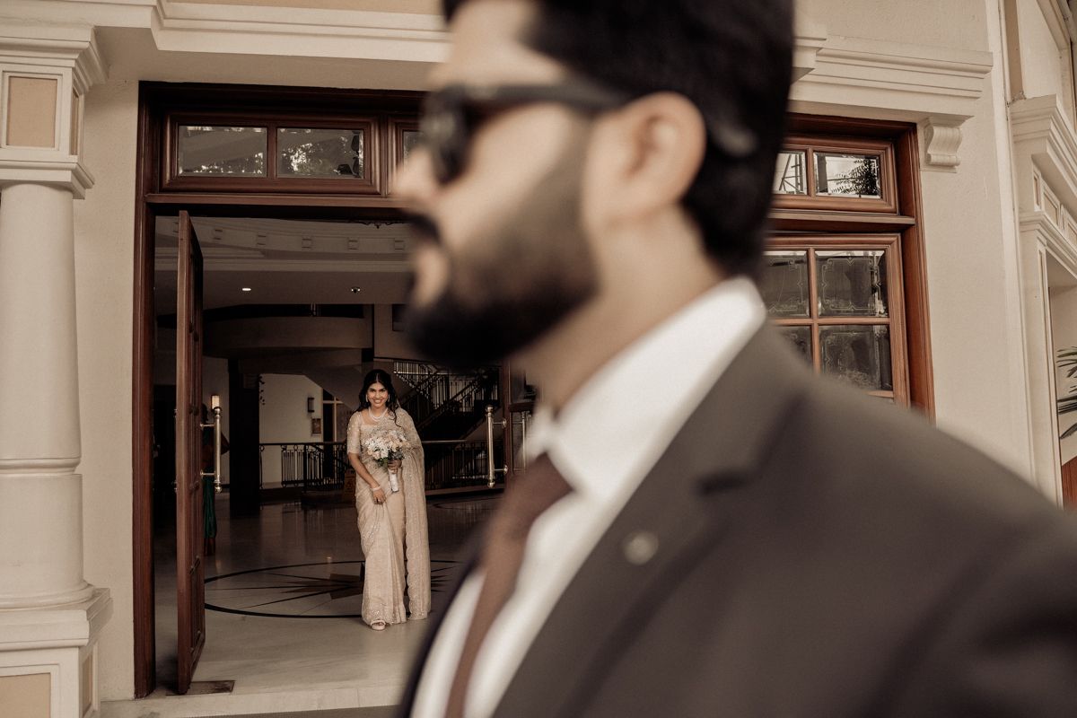 A sepia-toned wedding photograph with two figures standing at an elegant entrance with traditional architectural details.