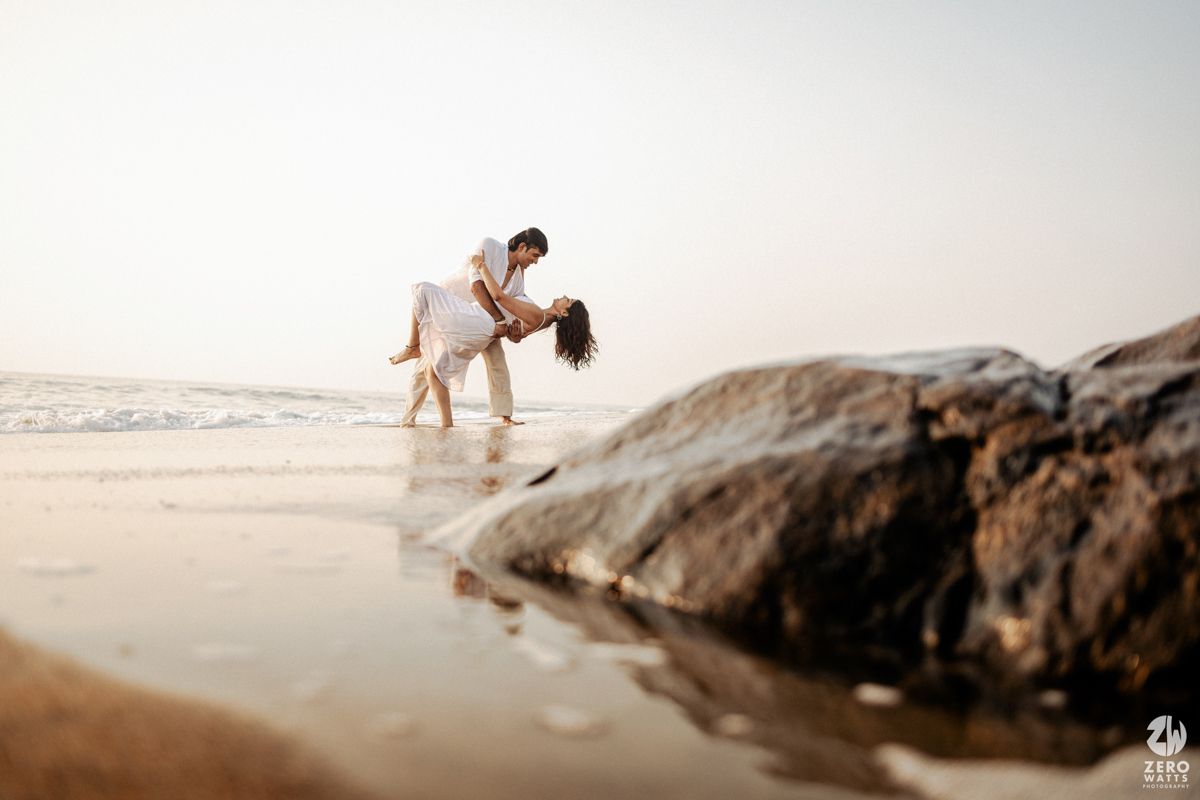 Couple dances romantically on beach with dramatic rock formations and ocean waves.