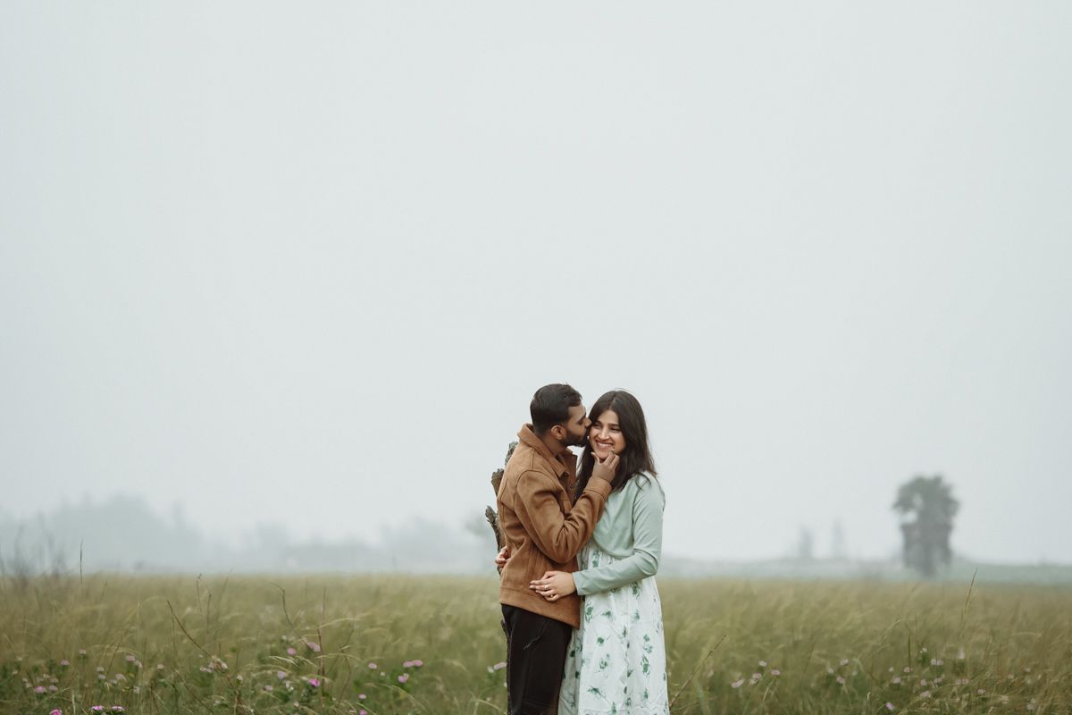 A couple stands close together in a misty field with long grass on an overcast day.