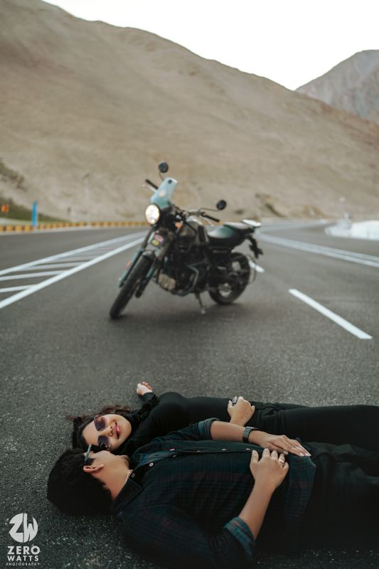 Person lying on asphalt road with motorcycle in background against desert mountain landscape.