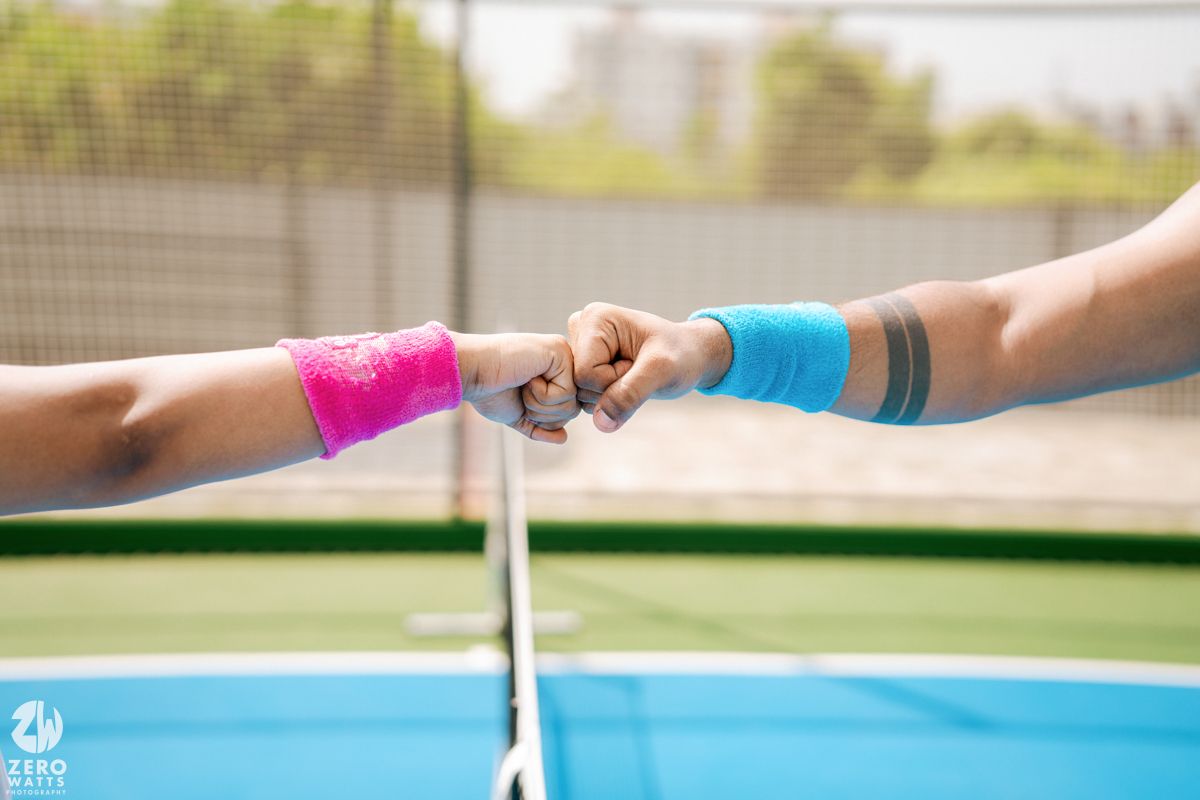 Close up of hands wearing pink and blue wristbands touching near a tennis net.