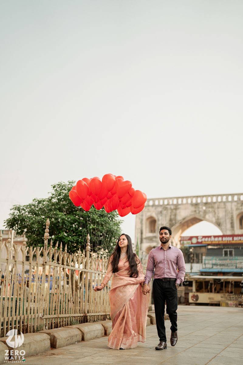 A romantic couple walks along a boardwalk carrying red heart balloons against a hazy sky.