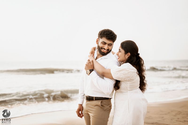 A romantic couple dancing together on a misty beach wearing white outfits during a photoshoot.
