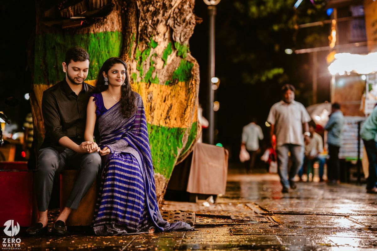 A nighttime street scene with couple on bench and pedestrians walking in background.